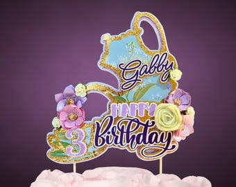 May include: A cake topper with the words "Gabby Happy Birthday 3" in a cursive font, with a gold outline and floral accents. The topper is on a pink frosted cake.