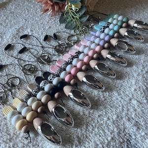 May include: A selection of pacifier clips in a variety of colours and patterns. Each clip has a string of beads, including solid colours, animal prints, and wooden beads. The clips have a silver-coloured metal end and a loop for attaching to a dummy.