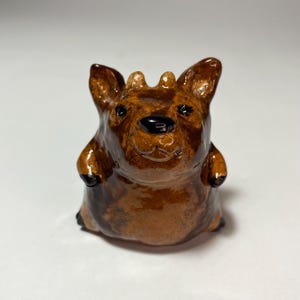 Könnte beinhalten: Eine kleine, handgeformte braune Keramikfigur eines Wesens mit spitzen Ohren, zwei kleinen hornartigen Auswüchsen auf dem Kopf und einer glänzenden Oberfläche. Die Figur hat einen abgerundeten Körper, dunkle Augen und eine schwarze Nase.