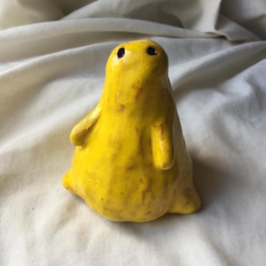 Puede incluir: Figurita de cerámica amarilla hecha a mano con cuerpo redondeado, brazos pequeños y dos puntos negros en forma de ojos. La escultura tiene una superficie texturizada y está sobre un fondo de tela blanca. Es un objeto decorativo único.
