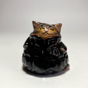 Puede incluir: Una pequeña figura de cerámica de un gato atigrado marrón con ojos amarillos y nariz rosa, que lleva una chaqueta acolchada negra brillante. El gato tiene un cuerpo redondeado y robusto, expresión facial neutra y un acabado de esmalte liso.