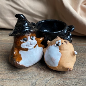 Puede incluir: Dos pequeñas figuras de gato de cerámica y un caldero de cerámica negra. Un gato es marrón y blanco con un sombrero puntiagudo negro, mientras que el otro es de color tostado claro y blanco con un sombrero negro más pequeño. Caldero negro brillante.