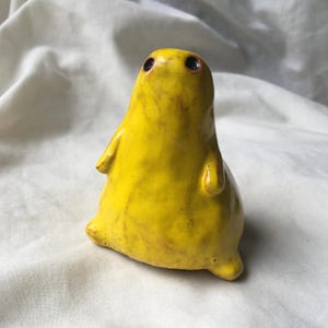 Op de afbeelding: Een klein, met de hand gevormd keramieken beeldje met een glanzend, felgeel glazuur. Het wezen heeft een afgerond, peervormig lichaam, twee kleine uitstekende armpjes en twee donkere, cirkelvormige inkepingen als ogen op de gladde kop.