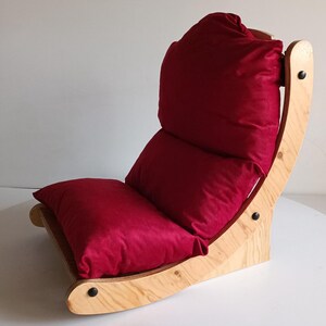 May include: A floor chair with a wooden frame and a deep red cushion. The chair has a curved design with a padded back and seat. The cushion is a solid color and appears to be made of a soft material. The chair is designed for comfort and relaxation.