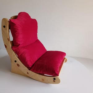 May include: A wooden rocking chair with a bright red cushion and backrest. The chair has a curved design with a natural wood finish. The seat and backrest are made of a soft, plush material.