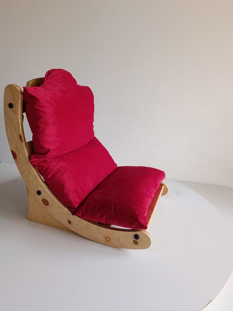 May include: A wooden rocking chair with a bright red cushion and backrest. The chair has a curved design with a natural wood finish. The seat and backrest are made of a soft, plush material.