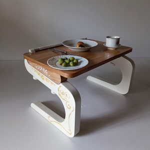 Może przedstawiać: Biały drewniany stolik z tacą z brązowego drewna. Stolik ma unikalny design z zakrzywioną podstawą i ozdobnymi detalami. Taca jest zastawiona talerzem oliwek, talerzem z ciastkiem oraz filiżanką i spodkiem.