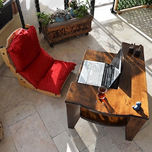 May include: An outdoor patio scene featuring a wooden coffee table with a laptop and a glass of tea. A red cushioned lounge chair and a woven basket are also present. Decorative planters with greenery add a natural touch.