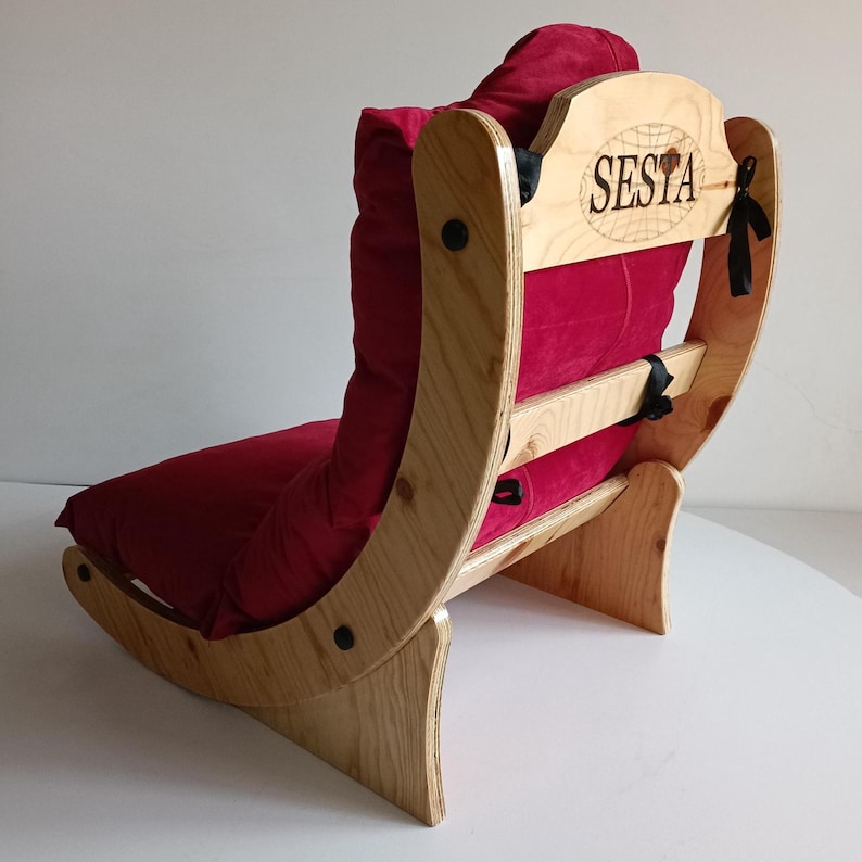 May include: A wooden lounge chair with a curved frame and a deep red cushion. The chair has a headrest with the word "SESTA" and a globe design. The frame is made of light-colored wood, and the cushion appears soft and inviting.
