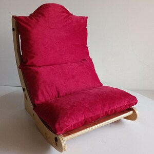 May include: A floor chair with a wooden frame and a plush, red cushion. The chair has a curved back and a low profile, suitable for sitting on the floor. The cushion is a deep red color, and the wooden frame is light-colored.