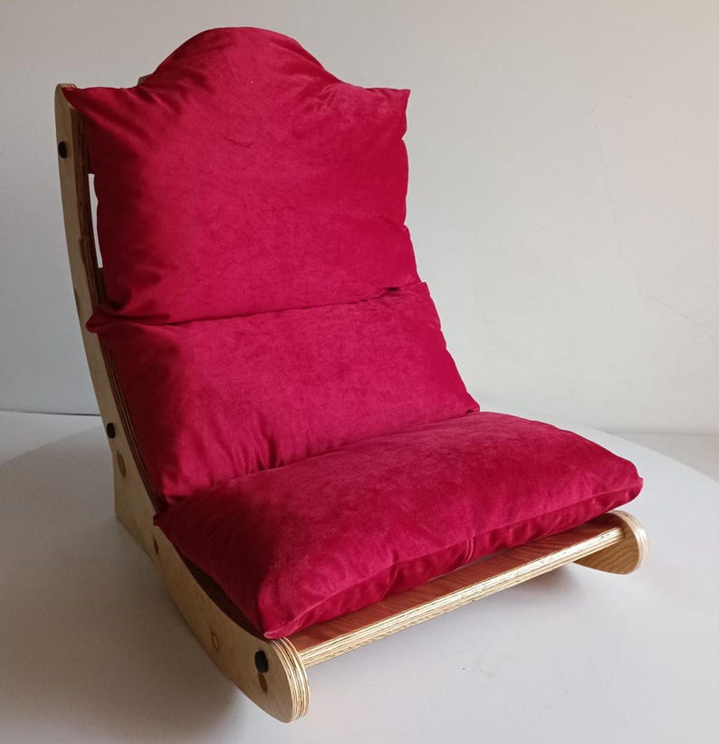 May include: A floor chair with a wooden frame and a plush, red cushion. The chair has a curved back and a low profile, suitable for sitting on the floor. The cushion is a deep red color, and the wooden frame is light-colored.