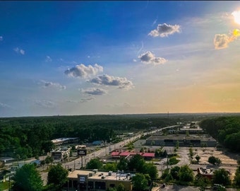 Può includere: Vista aerea di una zona suburbana con un ampio parcheggio e alcuni edifici. Il cielo è blu con nuvole bianche e il sole splende.
