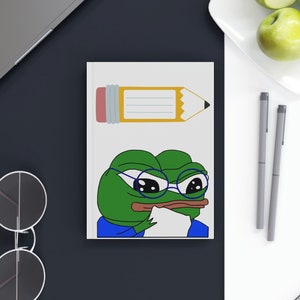 May include: A white notebook with a green frog wearing glasses and a blue shirt. The frog is thinking and has a pencil with a yellow eraser on the notebook. The notebook is on a black surface.