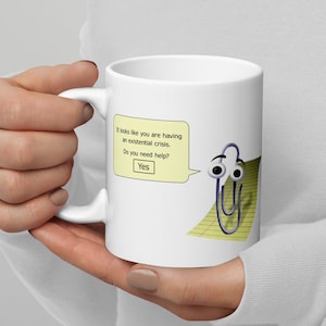 Könnte beinhalten: Eine weiße Keramiktasse mit einer blauen Büroklammer mit Glitzern und einer Sprechblase, die sagt: "Es sieht so aus, als hättest du eine existenzielle Krise. Brauchst du Hilfe? Ja".