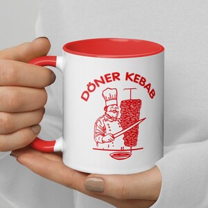 Könnte beinhalten: Weiße Keramiktasse mit rotem Rand und Henkel. Die Tasse zeigt eine rote Illustration eines Kochs, der einen Döner Kebab zubereitet, mit dem Text "Döner Kebab" in rot.