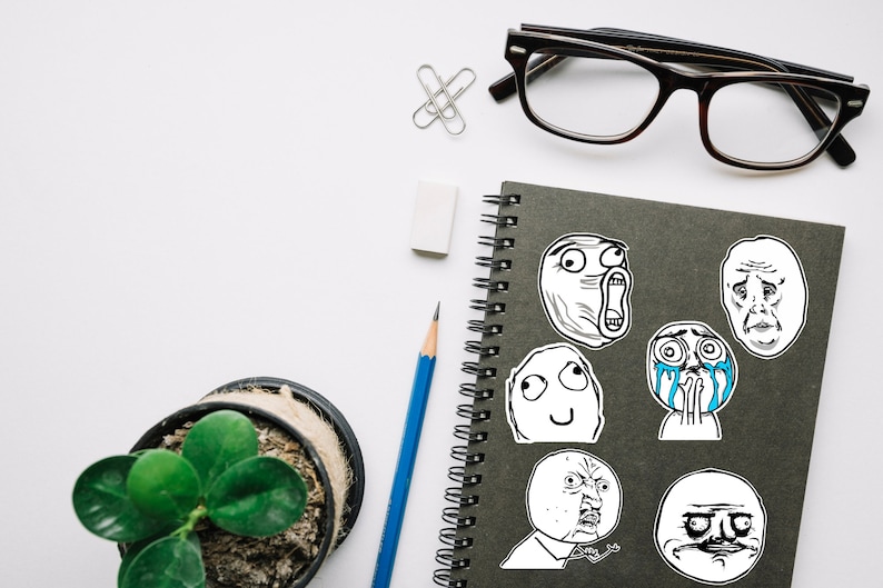May include: A black notebook with a spiral binding features several internet meme stickers. The stickers are black and white and depict various facial expressions. The notebook is on a white surface with a blue pencil, a white eraser, and a pair of black eyeglasses.