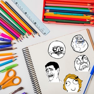 May include: A white notebook with black spiral binding, filled with black and white drawings of popular internet memes.  The notebook is surrounded by colored pencils, pens, and scissors on a white surface.