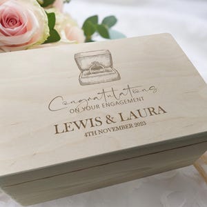 May include: A light-coloured wooden box with a hinged lid. The lid is engraved with a drawing of a ring in a box, the words "Congratulations on your engagement", and the names "LEWIS & LAURA" with the date "4TH NOVEMBER 2023".
