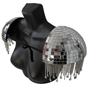 Könnte beinhalten: Silberne Disco-Kugel-Schulterpolster mit Fransen. Die Schulterpolster bestehen aus Spiegelkacheln und haben einen schwarzen Gurt, der sich auf der Rückseite binden lässt.