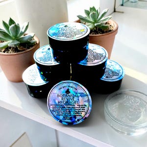 Puede incluir: Una pila de frascos de vidrio oscuro que contienen Botanical Revive Hydragel, con etiquetas iridiscentes que enumeran ingredientes como ácido hialurónico, hidrosol de rosa y gel de aloe vera. Un frasco transparente abierto se sitúa al lado.