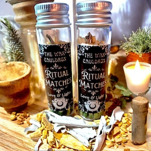 May include: Two glass bottles with silver lids, labeled "The Wax Cauldron Ritual Matches Love & Light." The bottles are filled with matches and sit on a wooden surface with herbs and a lit candle.