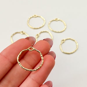 以下が含まれることがあります： 金色のテクスチャ加工されたオープンサークルチャームのクローズアップ。1つのチャームが手に持たれ、他のチャームは白い表面に散らばっています。各チャームには、他のジュエリーコンポーネントに取り付けるための小さなループが付いています。