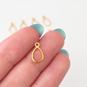 May include: Gold teardrop shaped pendant with a small loop at the top. The pendant is held in a hand.