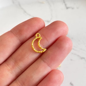 Peut inclure: Un petit charm en forme de croissant de lune doré avec une surface texturée. Le charm possède une boucle en haut pour l'attacher à un bijou. Le charm est probablement en métal et est conçu pour la fabrication de bijoux ou comme élément décoratif.
