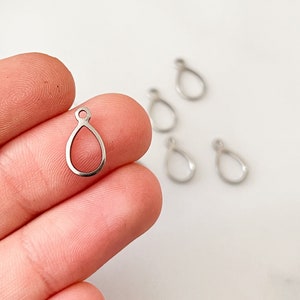 Könnte beinhalten: Silberne tropfenförmige Metall-Anhänger mit einer Öse oben. Die Anhänger sind auf einer Hand vor weißem Hintergrund zu sehen.