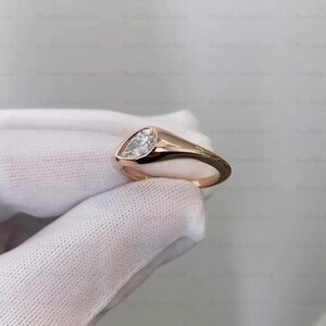 Puede incluir: Un anillo de oro rosa con un diamante en forma de pera. El anillo tiene una banda lisa y pulida y un engaste de bisel que sujeta el diamante de forma segura. El diamante es claro y brillante, y el anillo se muestra sobre un fondo blanco.