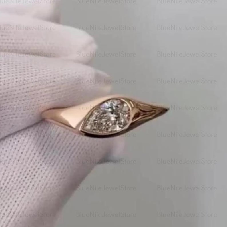 Puede incluir: Un anillo de oro rosa con una gema clara en forma de pera. El anillo tiene una banda lisa y pulida que se ensancha para acunar la piedra. La gema brilla, captando la luz. El anillo se muestra sobre un fondo suave y neutro.