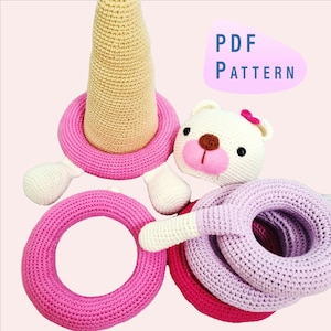 May include: A collection of handmade crochet baby toys in various shades of pink, white, and beige. The toys include a bear head, rings, and a cone-shaped element. The image also includes the text "PDF PATTERN".