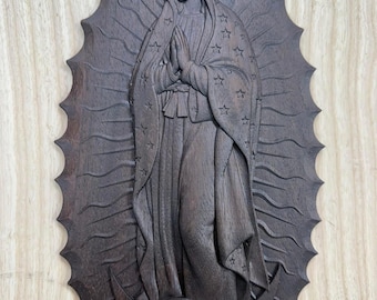 May include: A dark brown wooden carving of the Virgin Mary, hands clasped in prayer, wearing a star-studded cloak. The figure is set against a sunburst-shaped background with a detailed, carved design. The carving is mounted on a light-colored wooden surface.
