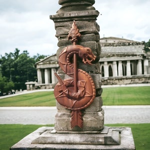 May include: A carved wooden wall hanging featuring a snake coiled around a straight arrow. The snake is depicted with an open mouth and textured scales, while the arrow has a pointed tip at the top and fletching at the bottom, all in brown.