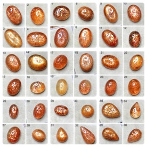May include: A collection of thirty-six loose sunstone gemstones arranged in a grid, each numbered and featuring varying shades of orange, copper, and gold with internal sparkling inclusions and smooth, polished surfaces in oval and teardrop shapes.