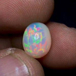 May include: A close-up of an oval-shaped opal gemstone. The opal has a milky white base color with vibrant flashes of green, blue, orange, and pink. The gemstone is held between two fingers, showcasing its iridescent play-of-color.