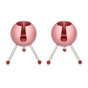 May include: Two pink metal candle holders with three legs each. The candle holders have a round top with a hole in the centre for a candle.