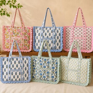 May include: A collection of six quilted tote bags with floral and leaf patterns. The bags feature ruffled edges and matching handles. Colors include pink, blue, and green, with white backgrounds. The bags are arranged on a wooden surface.