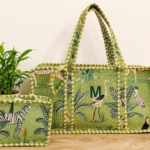 Puede incluir: Un bolso tote acolchado verde y un estuche rectangular a juego con estampado de selva, cebras, flamencos y follaje tropical. El bolso tiene dos asas y una letra M verde oscuro bordada. Ambos art&iacute;culos tienen bordes a rayas.