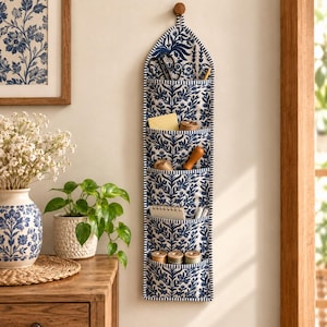 Könnte beinhalten: Ein vertikaler Wand-Organizer aus Stoff mit blau-weißem Blumenmuster und fünf gestuften Taschen. Der Organizer hängt an einem kleinen Holzknopf und enthält verschiedene kleine Gegenstände wie eine Schere, Garnrollen und einen Notizblock.