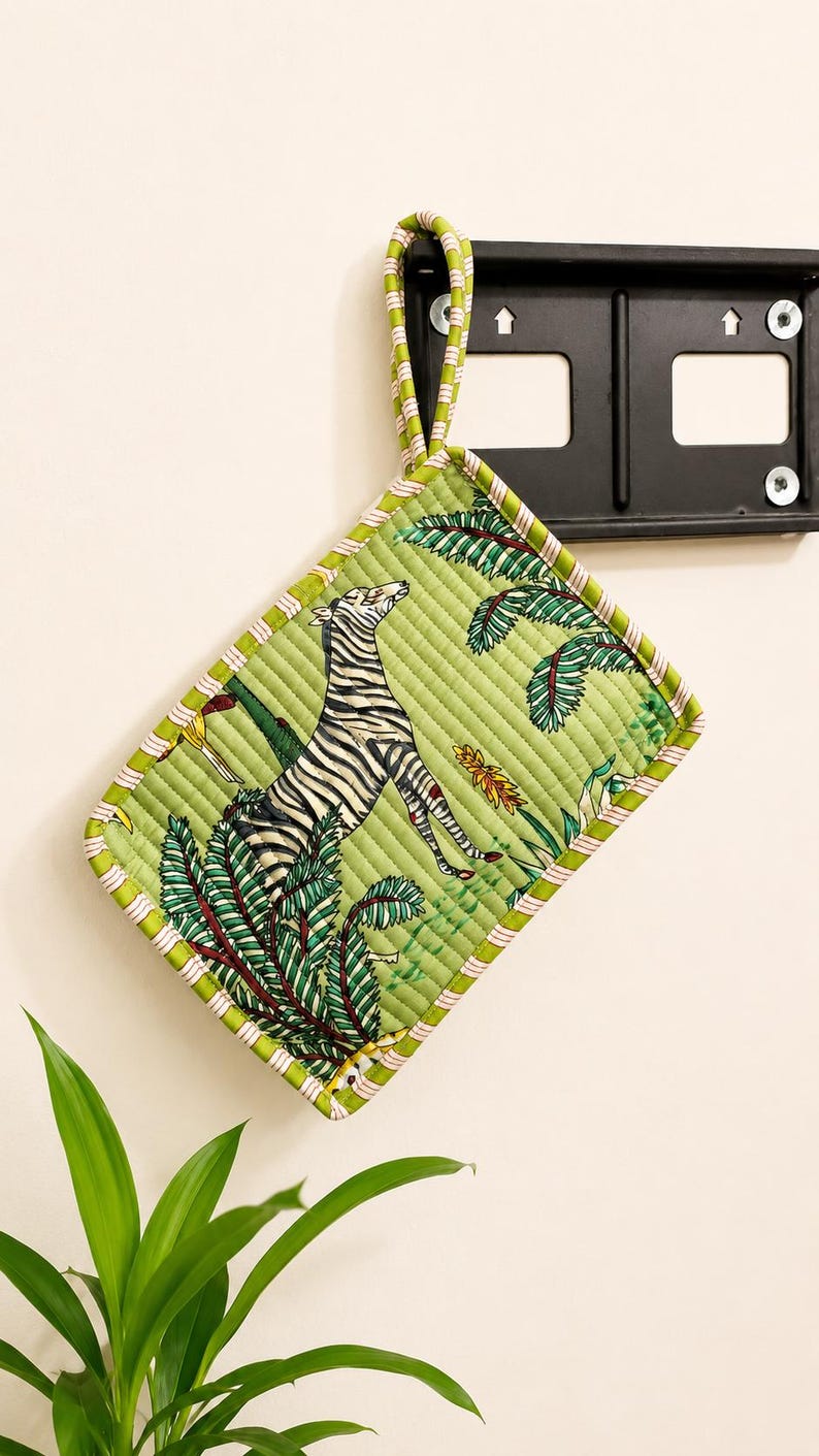 Puede incluir: Un agarrador de cocina acolchado rectangular con fondo verde claro y un patr&oacute;n estampado de una cebra y hojas de palmera tropicales. Los bordes est&aacute;n rematados con un ribete de tela a rayas verdes y blancas y una trabilla a juego para colgar.