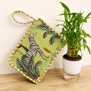 Puede incluir: Un estuche acolchado rectangular con fondo verde y un estampado de cebra y follaje tropical. El estuche incluye una correa de mu&ntilde;eca de tela a juego y un borde a rayas, mostrando una textura cosida detallada en toda la superficie.