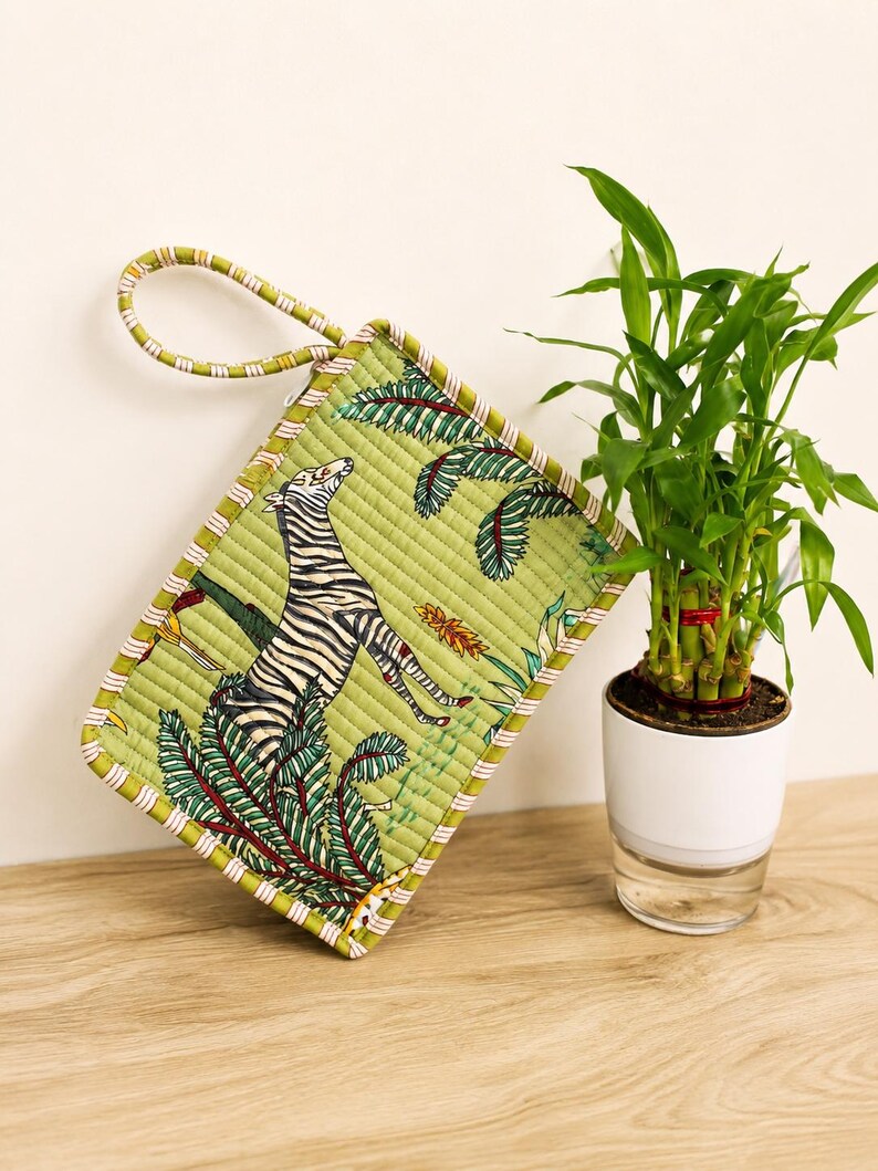 Puede incluir: Un estuche acolchado rectangular con fondo verde y un estampado de cebra y follaje tropical. El estuche incluye una correa de mu&ntilde;eca de tela a juego y un borde a rayas, mostrando una textura cosida detallada en toda la superficie.