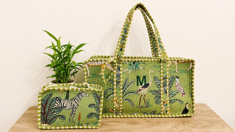 Puede incluir: Un bolso tote acolchado verde y un estuche rectangular a juego con estampado de selva, cebras, flamencos y follaje tropical. El bolso tiene dos asas y una letra M verde oscuro bordada. Ambos art&iacute;culos tienen bordes a rayas.