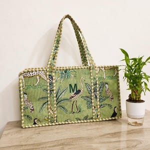 Puede incluir: Un bolso tote acolchado rectangular de color verde claro con un patr&oacute;n repetitivo de aves tropicales, guepardos y hojas de palmera. El bolso tiene dos asas a juego con rayas verdes y blancas y una letra M bordada en verde oscuro.