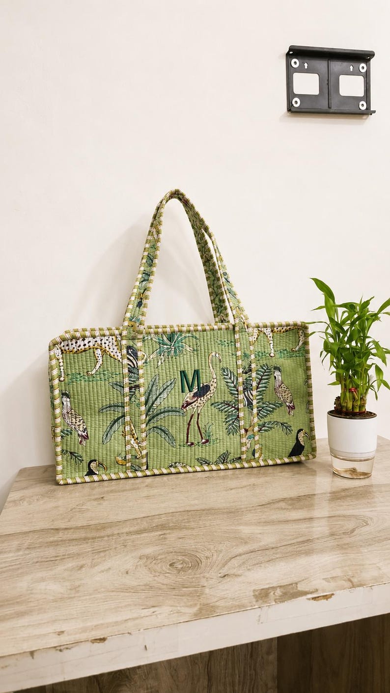 Puede incluir: Un bolso tote acolchado rectangular de color verde claro con un patr&oacute;n repetitivo de aves tropicales, guepardos y hojas de palmera. El bolso tiene dos asas a juego con rayas verdes y blancas y una letra M bordada en verde oscuro.