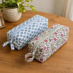 K&ouml;nnte beinhalten: Zwei rechteckige Federm&auml;ppchen mit floralen Mustern. Eines ist blau mit wei&szlig;en Blumen, das andere rosa mit wei&szlig;en Blumen. Beide haben Rei&szlig;verschl&uuml;sse und Stoffschlaufen. Die Etuis liegen auf einer Holzoberfl&auml;che.