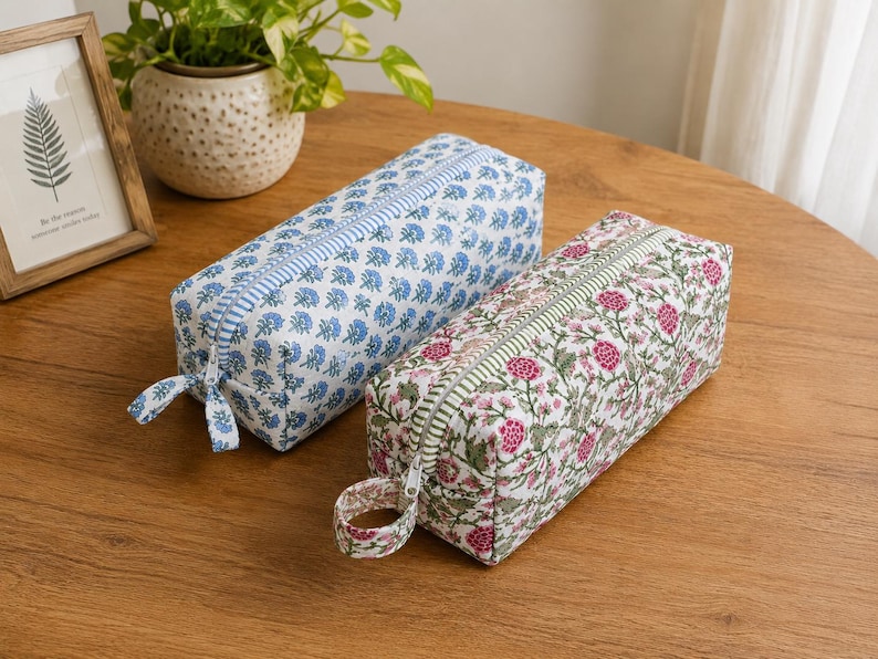 K&ouml;nnte beinhalten: Zwei rechteckige Federm&auml;ppchen mit floralen Mustern. Eines ist blau mit wei&szlig;en Blumen, das andere rosa mit wei&szlig;en Blumen. Beide haben Rei&szlig;verschl&uuml;sse und Stoffschlaufen. Die Etuis liegen auf einer Holzoberfl&auml;che.