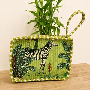 Puede incluir: Una cesta rectangular de tela acolchada con fondo verde y un estampado de cebra y follaje tropical. La cesta tiene un asa arqueada &uacute;nica y un borde a rayas, y contiene una peque&ntilde;a planta verde de hojas.