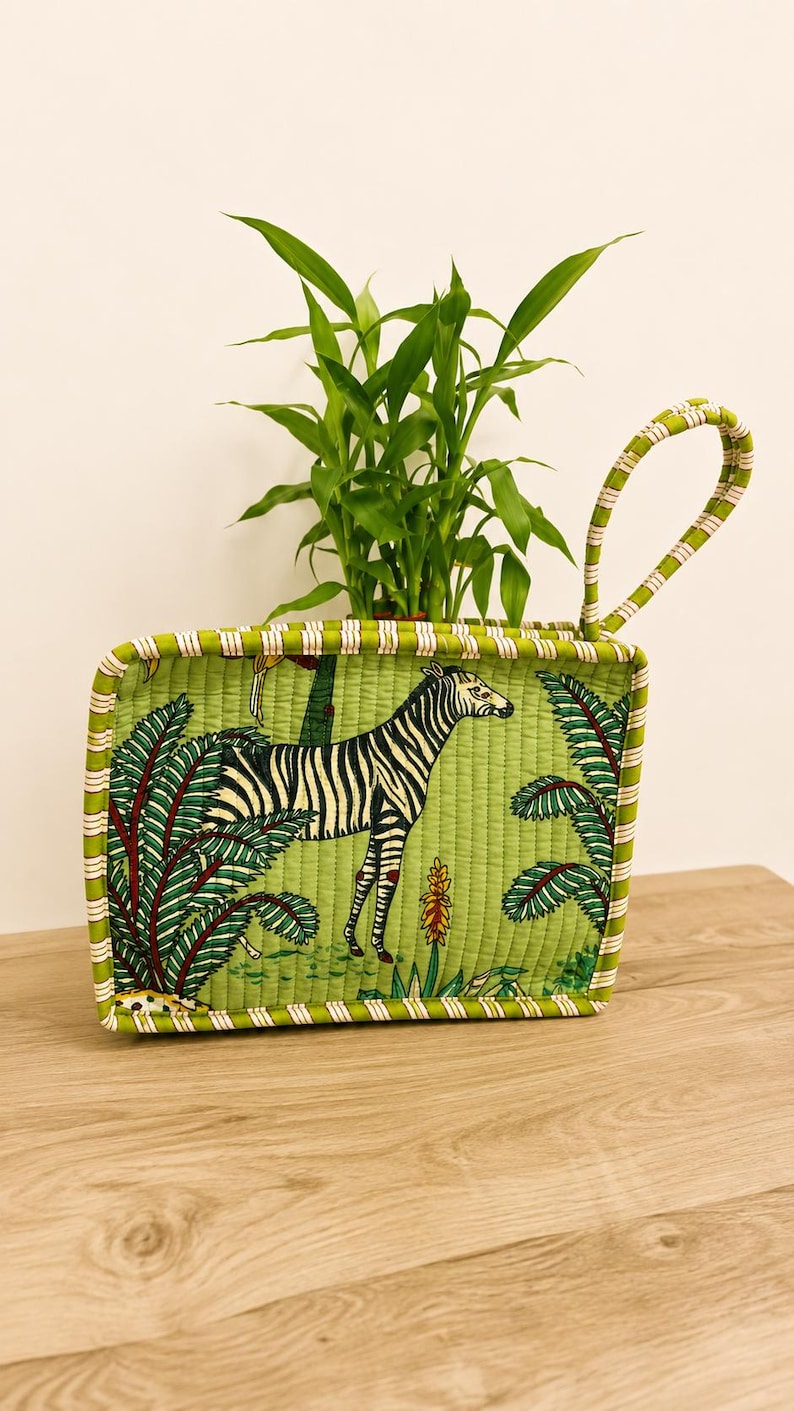 Puede incluir: Una cesta rectangular de tela acolchada con fondo verde y un estampado de cebra y follaje tropical. La cesta tiene un asa arqueada &uacute;nica y un borde a rayas, y contiene una peque&ntilde;a planta verde de hojas.